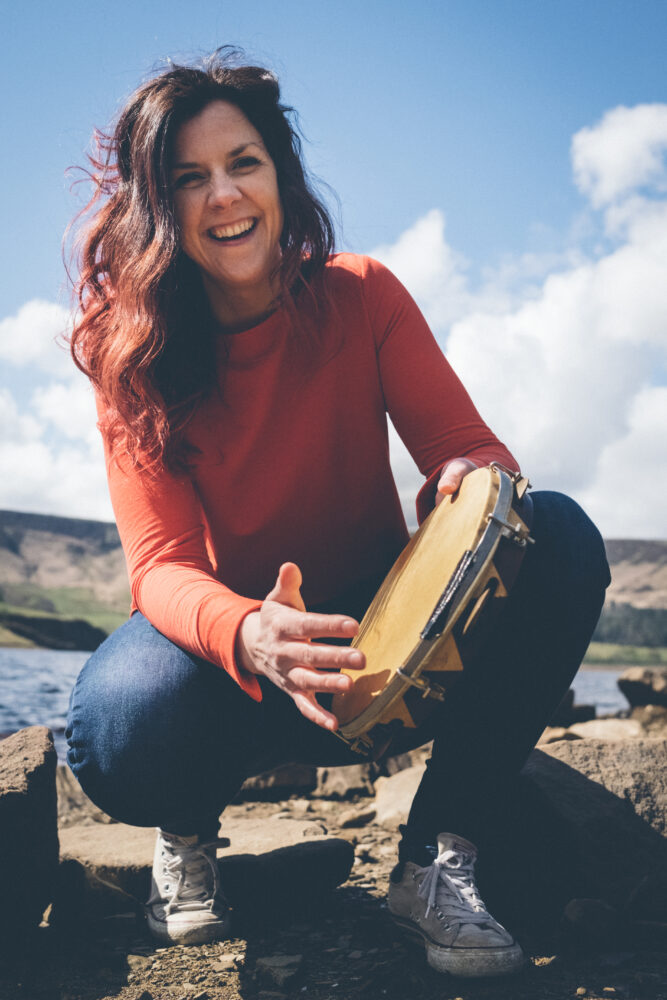 Percussionist Holly Prest kneeling down playing a pander drum on a sunny day